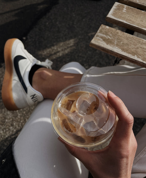 Hand holding iced coffee outdoors beside white Nike sneakers on
sunny day