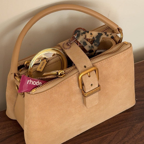 Tan suede mini handbag with gold hardware and Rhode lip product on
wooden surface